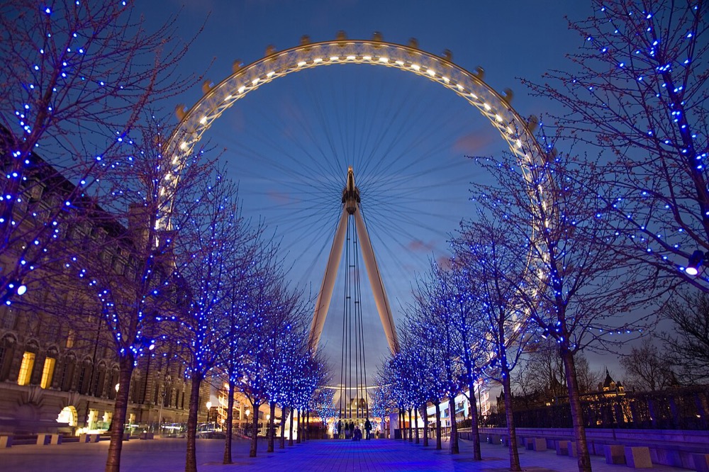 The London Eye lit at twilight, seen along an avenue of blue-lit trees